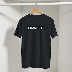 Puede incluir: Camiseta negra con la frase blanca "CHARGE IT.". La prenda está colgada de una percha de madera sobre un fondo neutro.