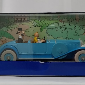 May include: A light blue vintage car toy with white tires and a detailed interior. Two figurines are inside the car, and the background shows a jungle scene. The toy is displayed in a blue box.