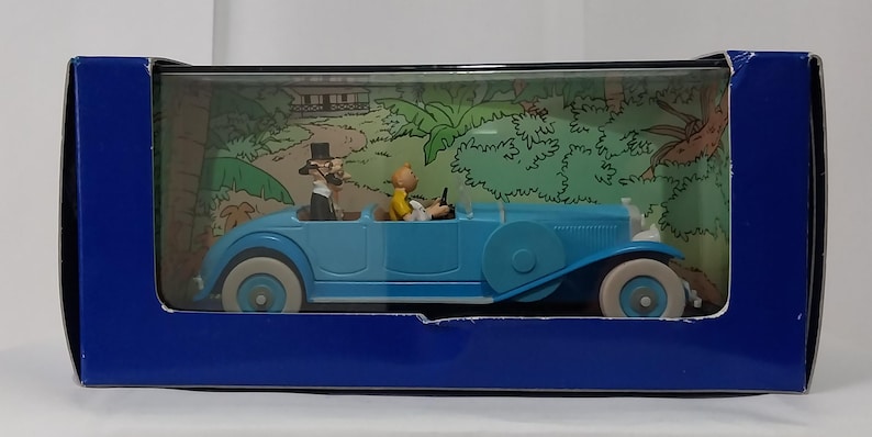 May include: A light blue vintage car toy with white tires and a detailed interior. Two figurines are inside the car, and the background shows a jungle scene. The toy is displayed in a blue box.