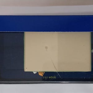 Puede incluir: Una caja rectangular azul con una ventana transparente, que revela un producto en su interior. La caja tiene un interior blanco y un exterior azul oscuro. El texto "CILE NOIRE" es visible en el producto.