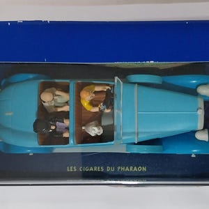 May include: A light blue vintage car model with figures inside, presented in a blue box. The car has a classic design with rounded fenders and a convertible top. The box has the text "LES CIGARES DU PHARAON".
