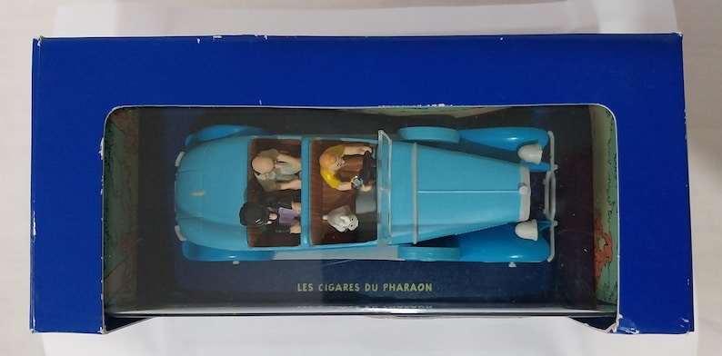 May include: A light blue vintage car model with figures inside, presented in a blue box. The car has a classic design with rounded fenders and a convertible top. The box has the text "LES CIGARES DU PHARAON".