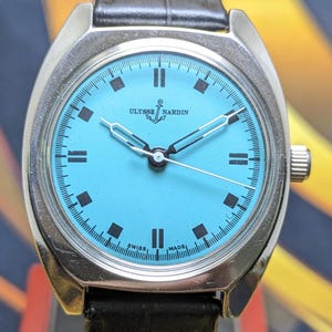May include: A silver Ulysse Nardin wristwatch with a turquoise face and black markers. The watch has a black leather strap and a silver bezel. The watch face displays the words "Ulysse Nardin" and "Swiss Made".