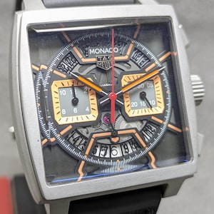 May include: A TAG Heuer Monaco watch with a square face, black and orange dial, and silver case. It has a black strap. The dial includes two sub-dials and a date display. This luxury watch is a stylish accessory.