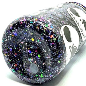 May include: A glitter-covered tumbler with a black base and a white skull design. The tumbler is covered in a mix of colorful glitter, including purple, green, and blue. The design features a skull and flame pattern.