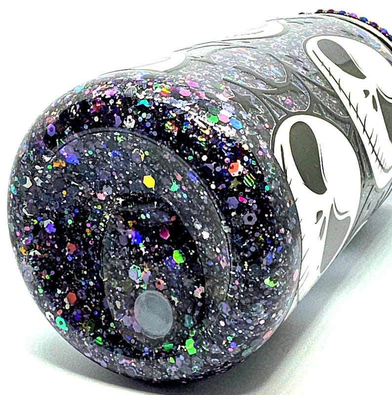 May include: A glitter-covered tumbler with a black base and a white skull design. The tumbler is covered in a mix of colorful glitter, including purple, green, and blue. The design features a skull and flame pattern.