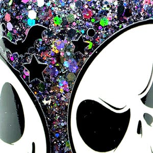 May include: A close-up of a Halloween-themed tumbler with a white skull design and a black bat and star design. The tumbler is filled with colorful glitter in various shapes and sizes, creating a sparkling effect.