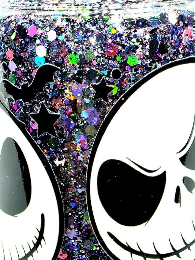 May include: A close-up of a Halloween-themed tumbler with a white skull design and a black bat and star design. The tumbler is filled with colorful glitter in various shapes and sizes, creating a sparkling effect.