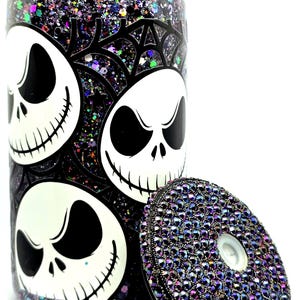 May include: A clear glass tumbler with a black and white Jack Skellington design and a bedazzled lid. The tumbler is filled with glitter in shades of purple, black, and iridescent colors. The lid is covered in small, iridescent rhinestones.