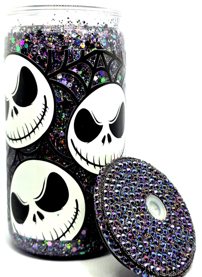 May include: A clear glass tumbler with a black and white Jack Skellington design and a bedazzled lid. The tumbler is filled with glitter in shades of purple, black, and iridescent colors. The lid is covered in small, iridescent rhinestones.