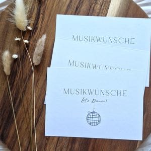 Könnte beinhalten: Drei weiße rechteckige Karten mit schwarzem Text MUSIKWÜNSCHE und einer kleinen Discokugel-Grafik mit dem Text Let's Dance auf der vorderen Karte. Die Karten liegen gestapelt auf einem runden Holzbrett neben getrockneten beigen Grashalmen.