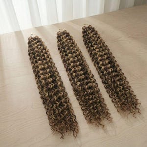 Puede incluir: Tres mechones de extensiones de cabello rizado marrón y rubio están dispuestos en diagonal sobre una superficie de madera clara. Cada mechón tiene un anillo dorado en la parte superior. El cabello tiene un aspecto natural y texturizado, con tonos de marrón y rubio.