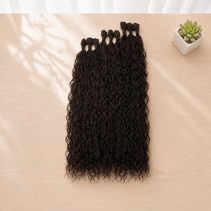 Puede incluir: Una colección de extensiones de cabello castaño oscuro con una textura de rizos apretados. Cada mechón individual está asegurado en la parte superior con un pequeño punto de unión de color oscuro, creando una fila uniforme de piezas de cabello ondulado.