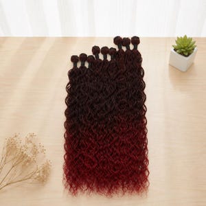 Puede incluir: Varios paquetes de extensiones de cabello ondulado, ombre burdeos oscuro y negro, están dispuestos sobre una superficie de madera clara. El cabello está muy rizado y pasa de oscuro a un tono rojo vibrante. Una pequeña planta en maceta está en el fondo.