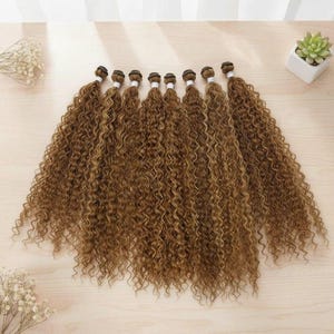 Peut inclure: Huit paquets d'extensions de cheveux bouclés dans une teinte brun doré chaud. Chaque paquet est fixé en haut par une bande blanche, présentant des boucles en tire-bouchon serrées et uniformes qui tombent en cascade.