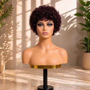 May include: A mannequin head with a short, dark burgundy wig featuring tight, voluminous curls. The wig has a rounded shape that frames the face, and the mannequin has a smooth, light-toned complexion with defined facial features and makeup.
