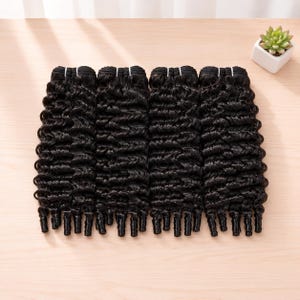 Può includere: Sei fasci di extension per capelli neri, molto ricci, sono disposti su una superficie di legno chiaro. I capelli sono di un nero intenso, con un motivo di ricci uniforme. Un piccolo vaso con una succulenta si trova nell'angolo in alto a destra.