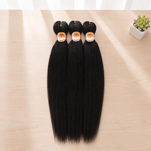 May include: Three bundles of straight, jet-black synthetic hair extensions arranged vertically. Each bundle is secured at the top with a circular paper band featuring a sunburst graphic and the text PRE-LAYERED in white and orange lettering.