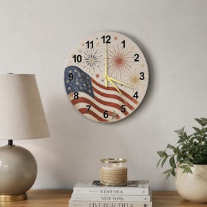 Puede incluir: Un reloj de pared redondo con un diseño patriótico. La esfera del reloj presenta la bandera estadounidense y fuegos artificiales. El reloj tiene números negros y manecillas amarillas. El reloj está colgado en una pared blanca.