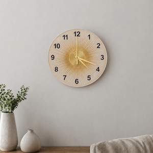 May include: A round wall clock with a gold sunburst design and black numerals. The clock hands are gold, and the clock is mounted on a light gray wall. The clock is approximately 12 inches in diameter.
