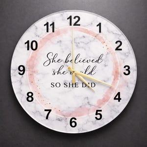 Puede incluir: Un reloj de pared redondo con un fondo similar al mármol y números negros. La esfera del reloj presenta un anillo rosa estilo acuarela y el texto "She believed she could SO SHE DID" en escritura negra. Las manecillas del reloj son doradas.