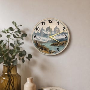 Op de afbeelding: Ronde wandklok met een landschap van bergen, een meer en een blauwe lucht. De wijzerplaat heeft zwarte cijfers en gouden wijzers. De klok is bevestigd aan een lichtgekleurde muur.