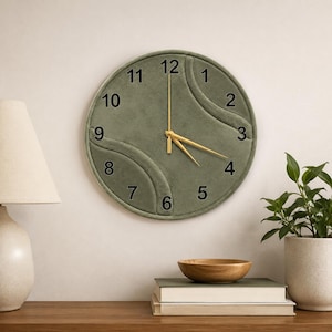 May include: A round wall clock with a sage green velvet-textured face featuring a raised, curved design element. The clock has black Arabic numerals from 1 to 12 and thin, gold-colored metal hands for the hour, minute, and second indicators.