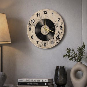 May include: Round wall clock with a vintage vinyl record design. The clock face features musical notes and the words "Music is life." Gold-colored hands indicate the time. The clock is mounted on a gray wall.