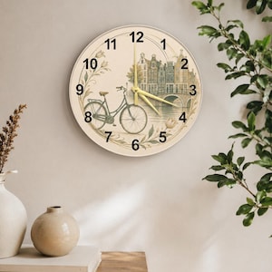 Könnte beinhalten: Runde Wanduhr mit cremefarbenem Zifferblatt, das eine Illustration von Fahrrad, Gebäuden und Brücke zeigt. Die Uhr hat schwarze Zahlen und Zeiger, mit einem goldenen Sekundenzeiger. Verziert mit floralen und Blättern. Durchmesser ca. 30 cm.