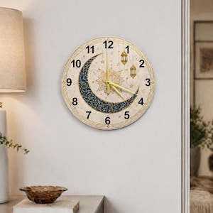 Puede incluir: Un reloj de pared redondo con una esfera color crema, con un diseño de media luna en azul marino y dorado. El reloj tiene manecillas doradas, números arábigos y elementos decorativos, incluyendo estrellas y faroles. Está colgado en una pared blanca.