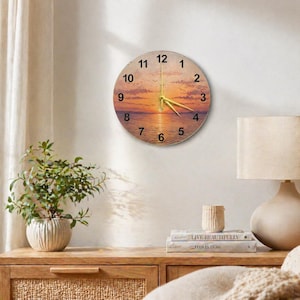 May include: Round wall clock featuring a sunset scene with a golden hour sky and water reflection. The clock has black numerals and gold hands. The clock is mounted on a white wall. The clock is approximately 12 inches in diameter.