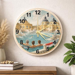 May include: Round wall clock with a cream-colored frame. The clock face features a painting of a thermal bath scene with people in the water and a Hungarian flag. Gold-colored hands indicate the time. Numbers are in black.