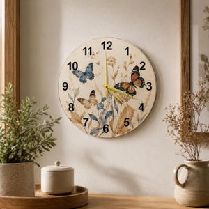 May include: A round wall clock with a cream-colored face featuring black numbers from 1 to 12. The clock face is decorated with illustrations of three butterflies in blue, orange, and brown, along with botanical sprigs. It has thin yellow hands.