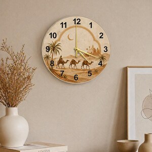 May include: Round wall clock with a desert scene featuring camels, palm trees, and a crescent moon. The clock face has gold-colored hands and Arabic numerals. The clock is mounted on a beige wall.