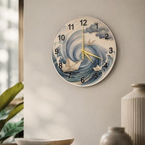 Könnte beinhalten: Eine runde Wanduhr mit einer kreisförmigen Illustration von blauen, wirbelnden Meereswellen und drei weißen Papierbooten. Das Zifferblatt zeigt schwarze arabische Ziffern von 1 bis 12 und drei dünne goldfarbene Zeiger.