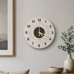 May include: A round wall clock with a cream-colored face. The clock features black numerals and a binary code design around the center. The clock hands are gold. The clock is hanging on a light gray wall.