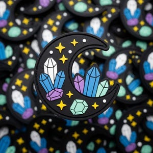 May include: Black crescent moon patch with colorful crystal and star designs. The crystals are white, blue, purple, and green. Yellow stars and white dots are scattered around the moon. The patch is likely made of rubber or a similar material.