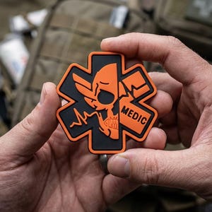 May include: A six-pointed star-shaped patch featuring a black background with an orange border and design. The design depicts a stylized skull split in half, an EKG heart rhythm line, and the word MEDIC printed in bold orange capital letters.