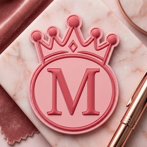 May include: A circular pink emblem featuring a raised letter M in the center, topped with a stylized crown design. The emblem has a smooth, matte finish with darker pink outlines and detailing, creating a layered appearance for the letter and crown.