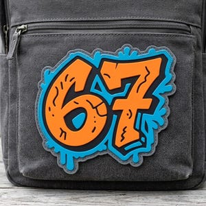May include: A charcoal gray canvas backpack featuring a large, vibrant patch on the front pocket. The patch displays the number 67 in bold orange with black outlines, set against a bright blue, dripping, graffiti-style background design.