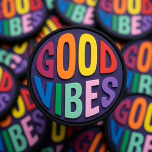 May include: Circular patch with the words "GOOD VIBES" in colorful, raised letters against a dark purple background. The letters are in rainbow colors, with a black border. Other similar patches are visible in the background.
