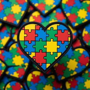 May include: A heart-shaped design featuring a puzzle pattern in red, yellow, green, and blue. The heart has a black border and is surrounded by similar designs. The puzzle pieces are interlocking, creating a colorful and symbolic image.