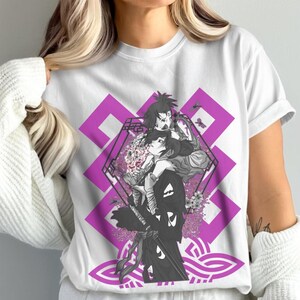 Op de afbeelding: Wit T-shirt met een afbeelding van twee anime-personages, een geometrisch paars ontwerp, bloemelementen en een zwaard. Het ontwerp is in grijstinten en paars. Het T-shirt heeft korte mouwen.