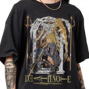 May include: Black t-shirt featuring a detailed anime graphic with the text "DEATH NOTE" in gold lettering. The design includes a blonde-haired character and other anime figures. The shirt has a vintage, worn look.