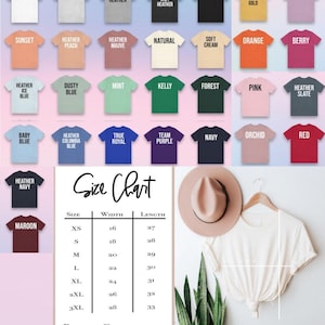 Op de afbeelding: Een verscheidenheid aan Bella + Canvas 3001 unisex jersey t-shirts in verschillende kleuren, waaronder wit, zwart en heather tinten. Een maattabel is inbegrepen, samen met een cr&egrave;mekleurige hoed en een plant.