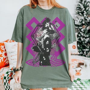 Op de afbeelding: Saliegroen t-shirt met een anime-achtige afbeelding van twee figuren, tegen een paars geometrisch ontwerp. De illustratie bevat bloemendetails en een gestileerd patroon. Het t-shirt heeft een losse pasvorm.