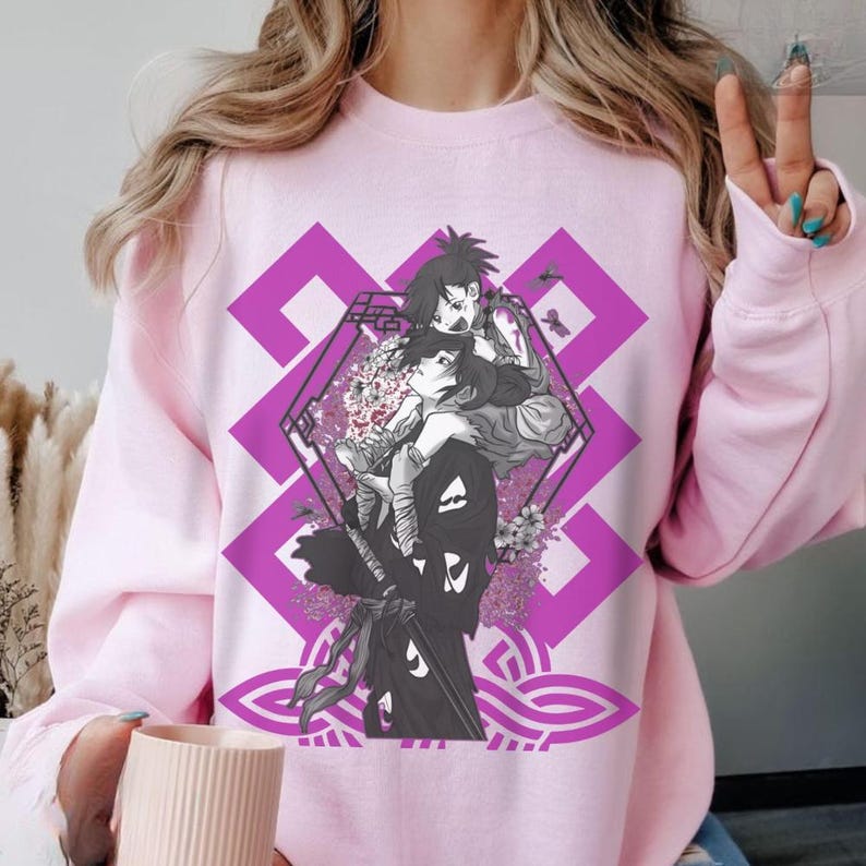 Op de afbeelding: Zachtroze sweatshirt met een afbeelding van twee anime-personages, geplaatst tegen een paars geometrisch en bloemenontwerp. De illustratie is gecentreerd op de voorkant van het kledingstuk.