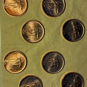 May include: Seven circular metal coins arranged in a grid pattern. Each coin features a raised relief design of a figure in profile, appearing to hold a long staff or spear. The coins exhibit varying shades of bronze, copper, and dark metallic tones.