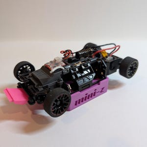 May include: A small, black and purple remote-controlled car with a pink front spoiler. The car has black wheels with red accents and visible internal components, including wires and a spring. The base of the car is purple and has the text "mini-z".