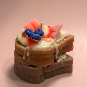 May include: A novelty candle shaped like a stack of two brown toast slices topped with white cream, red syrup, three blue berries, and two pink chocolate-like squares. A single white wick protrudes from the center of the cream topping.